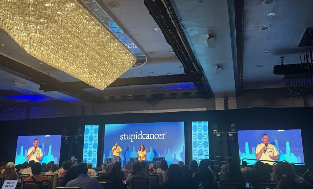 StupidCancer conference stage with LED screens, speakers, and presenters, showcasing event production by TriVision Studios in a large ballroom setting.