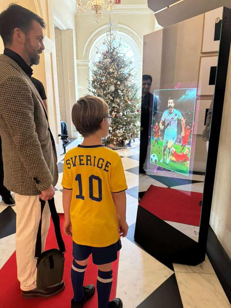 A boy and an adult engage with a holographic soccer player display at a festive event, with a decorated Christmas tree in the background.