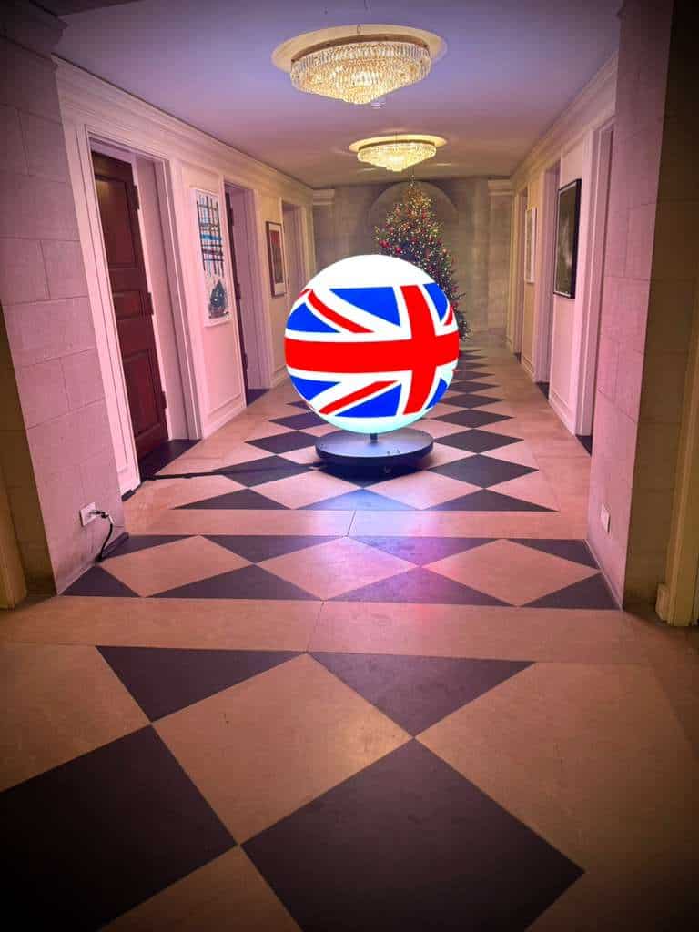A decorative LED globe featuring the Union Jack design is displayed in a luxury hotel hallway, accented by chandeliers and Christmas tree, showcasing modern lighting solutions.
