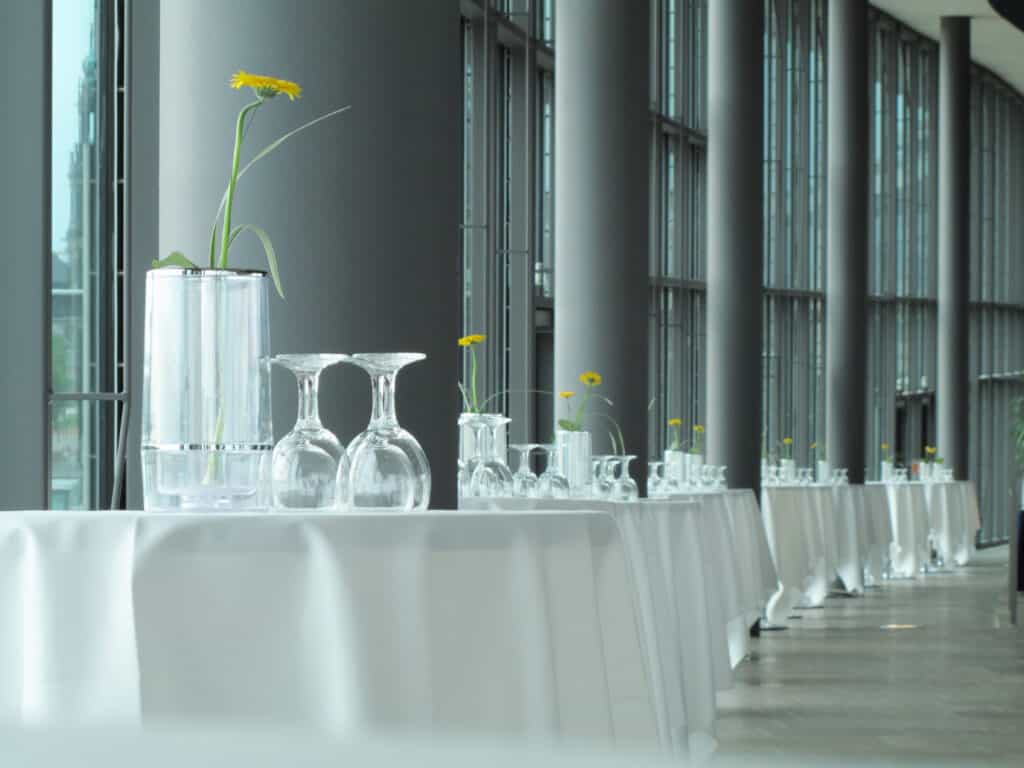 Modern indoor event space featuring a long table with glassware and small yellow flowers in vases, illuminated by natural light from large floor-to-ceiling windows, ideal for corporate gatherings or celebrations.
