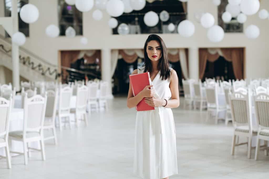 Stylish woman holding a folder at an upscale venue with white chairs and draped curtains.