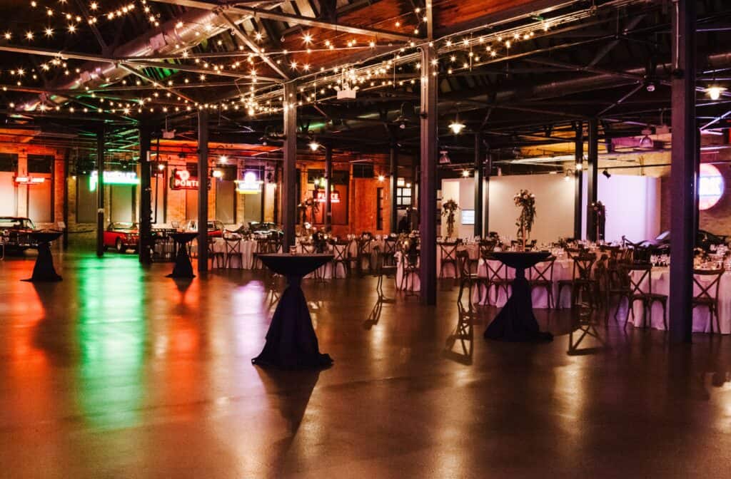 Brightly lit event space with string lights, vintage cars, and decorated tables for upscale parties or corporate events.