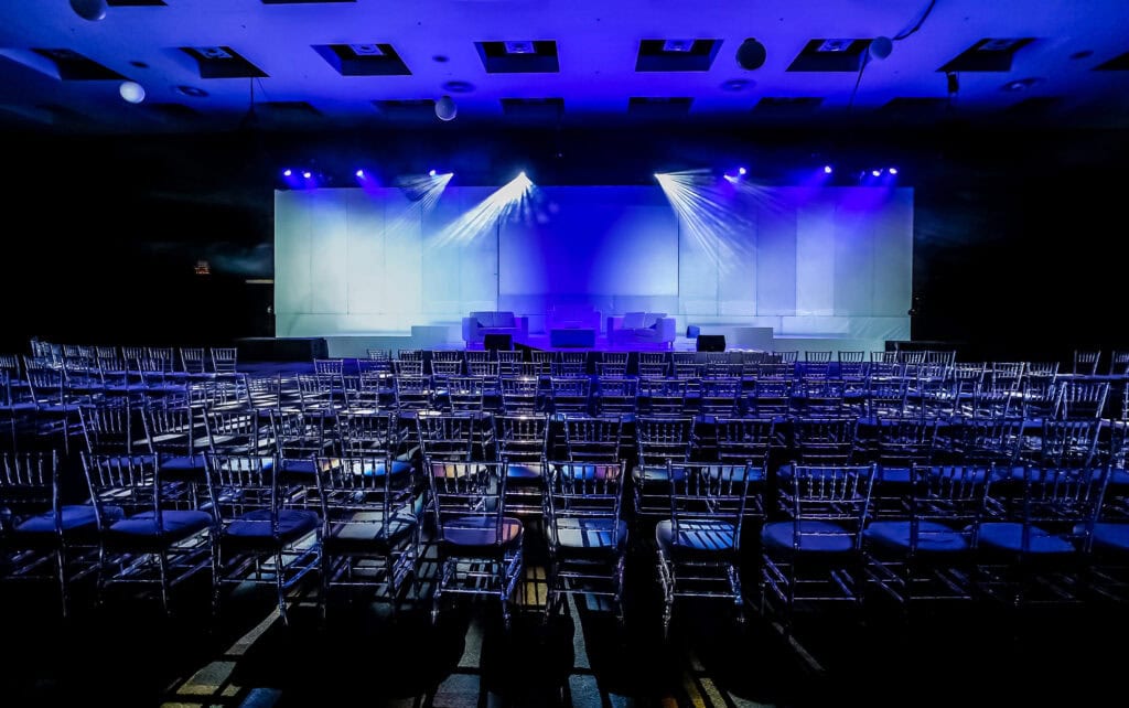 Bright stage lighting in a professional event venue with empty chairs for audience seating and a large white backdrop for presentations.