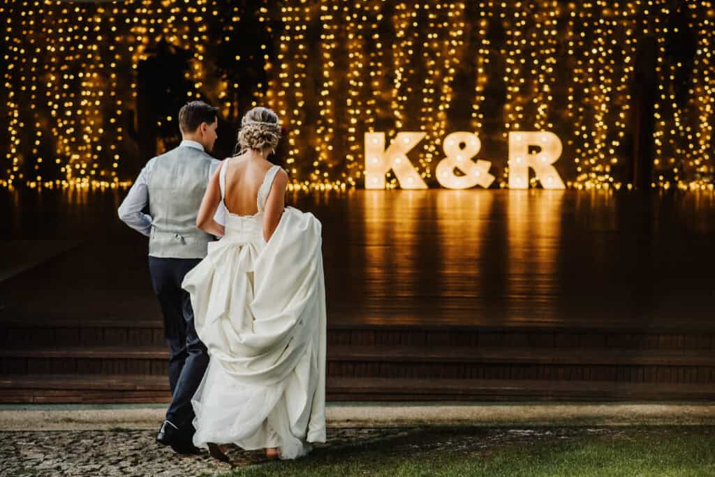 Beautiful newlywed bride and groom strolling outdoors during wedding reception with illuminated decorations and large light-up initials K & R.