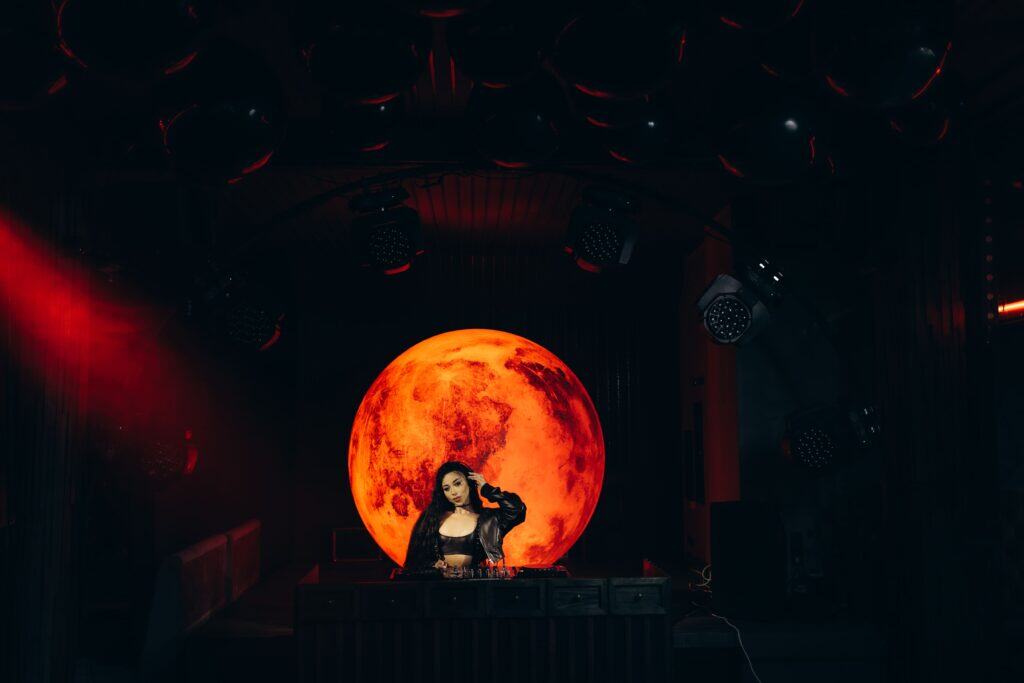 A female DJ performing on stage with a large, glowing orange moon backdrop creating a captivating atmosphere at a live event. Modern lighting and sound equipment enhance the energetic vibe.
