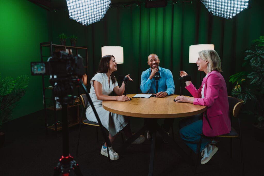 Interview recording with three diverse individuals seated around a table, engaged in conversation, with a camera capturing the scene in a well-lit, green-screen studio environment.
