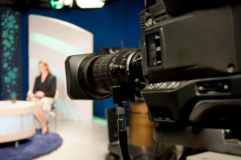 A high-quality professional video camera positioned in a TV studio, capturing a presenter seated on a set with a blue backdrop and a large screen behind her, ready for filming or broadcasting.